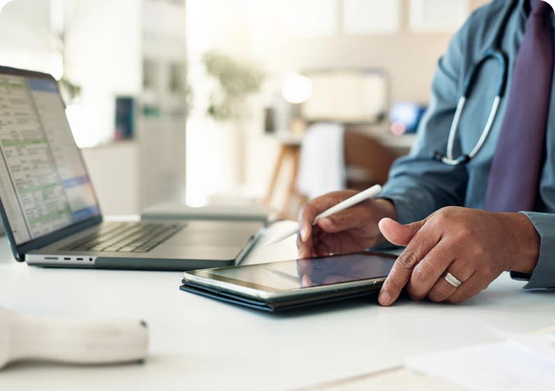 Doctor using tablet and laptop at desk.